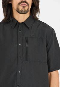 Zwarte korte mouwen shirt van gestructureerde stof, met een knoopsluiting aan de voorkant, een ritsportemonnee op de borst en een kraag.