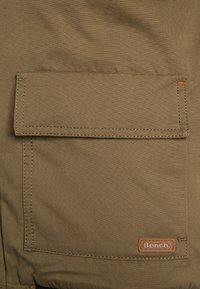 Kurtka oliwkowozielona z przednią kieszenią z klapką, przeszyciami i brązową naszywką z logo "Bench." Teksturowana powierzchnia materiału.