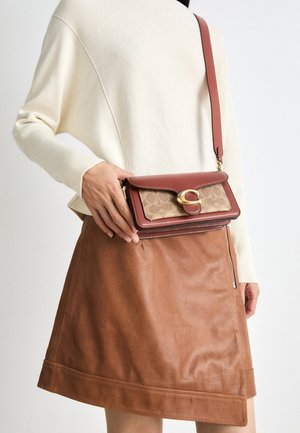 Woman wearing a cream sweater and brown leather skirt holding a brown and beige crossbody bag with a gold clasp.