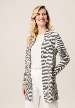 Blonde vrouw die lacht, draagt een wit topje en broek, en een lange zwart-witte jas met chevronpatroon, met één hand in de zak.