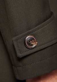 Zielono-oliwkowy materiałowy mankiet z kwadratowym wzorem, posiadający błyszczącym metalowym guzikiem z wygrawerowanymi detalami i teksturowaną powierzchnią.