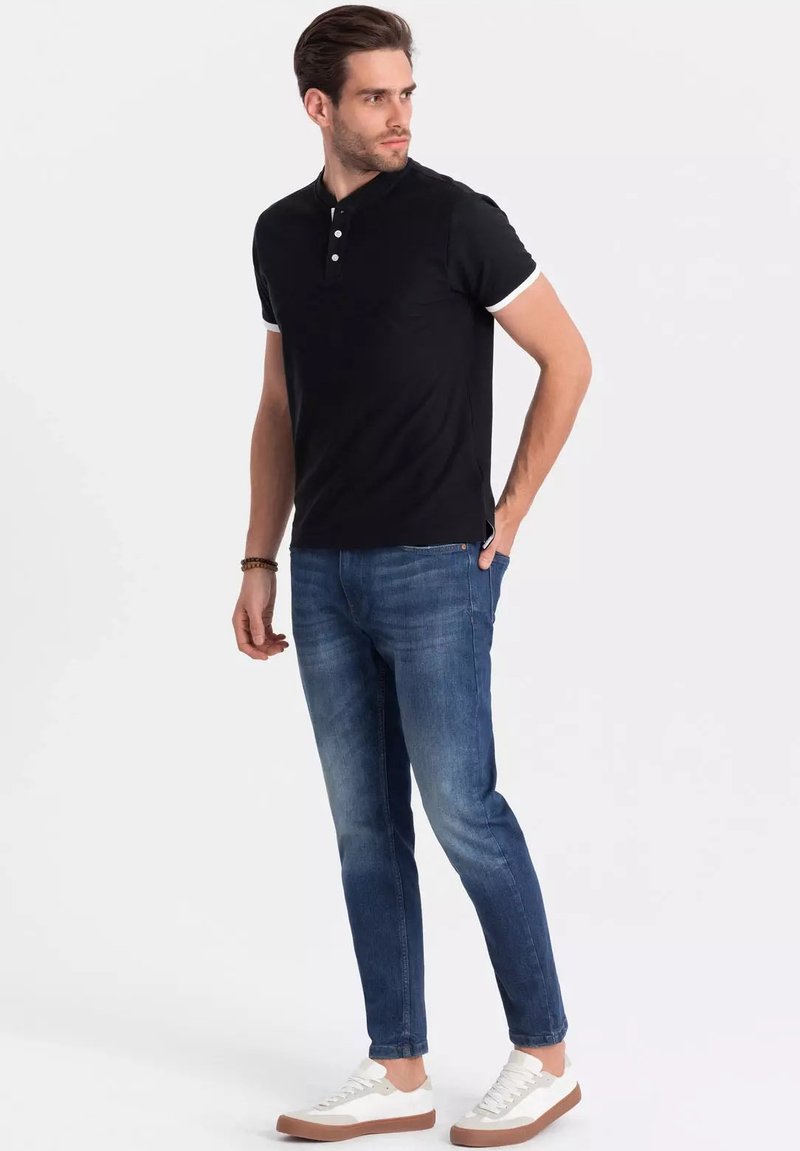 Černé polo tričko s bílými akcenty na límci, kombinované s modrými skinny džíny a bílými teniskami. Modelka stojí, ruce v kapsách.