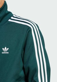 Tamnozeleni Adidas trenirku s visokim ovratnikom, bijelim trostrukim trakama na ramenima i bijelim logotipom na prsima.