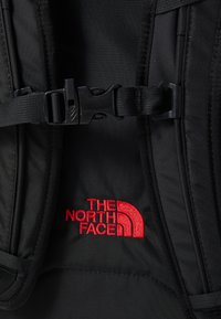 Svart sekk med slitesterkt stoff, justerbare stropper og en fremtredende rød brodert logo av 'The North Face' med et buet design.