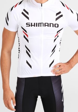 Cycliste masculin portant un maillot blanc Shimano avec des rayures noires et rouges et un cuissard noir assorti avec le logo Shimano.