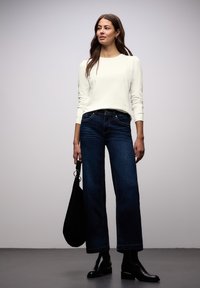 Wit top met lange mouwen en geplooide schouderdetails, gecombineerd met donkerblauwe jeans met wijde pijpen. Zwarte enkellaarsjes en een zwarte tas maken de look compleet.