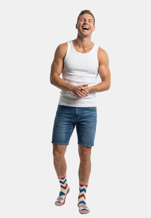 Weißes Tanktop, blaue Jeansshorts und mehrfarbige Socken mit Zickzackmuster. Die Person steht mit verschränkten Armen da und lächelt.