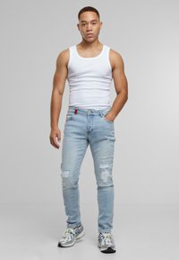 Light blue distressed jeans with rips, paired with a white tank top. Model wears gray and silver running shoes. Plain gray background.
