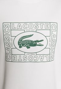 Logo crocodile vert Lacoste centré dans un cadre rectangulaire étiqueté "Lacoste Classics Since 1927" sur un fond en tissu blanc.