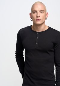 Crna dugosleeve henley majica od rebrastog materijala s četiri gumba, okruglim izrezom i prilagođenim krojem, nosi model na neutralnoj pozadini.