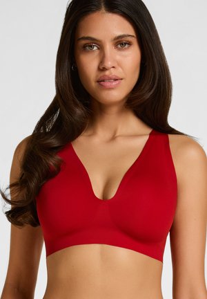 Vrouw met lang donker haar die een mouwloze rode bralette draagt tegen een effen lichte achtergrond.