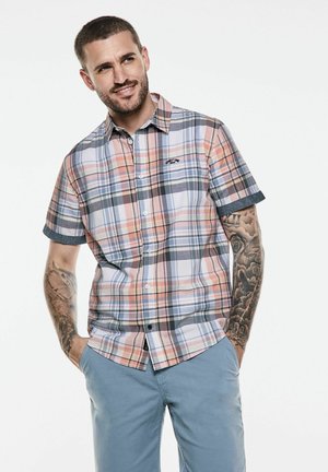 Chemise à manches courtes à carreaux avec des motifs rose, bleu et gris ; dotée d'une fermeture à boutons sur le devant et d'un logo sur la poche poitrine. Portée avec un pantalon bleu clair.