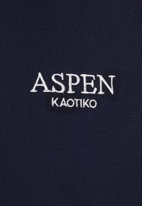 Mörkblått tyg med vit broderad text "ASPEN KAOTIKO." Slät yta utan synliga mönster eller hårdvarudetaljer.