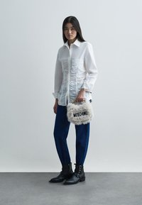 Balts, ručām apģērbs ar garām piedurknēm, zili bikses, melni potīšu zābaki ar sprādzēm; tur rokā pūkainu, krēmkrāsas somu ar melnu uzrakstu "MOSCHINO".