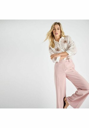 Blonde vrouw draagt een witte, bloemrijke blouse met knopen en een high-waist roze broek met wijde pijpen, poseert met gekruiste armen tegen een witte achtergrond.