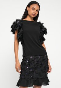 Fekete ruha, amely testhezálló felsőrésszel rendelkezik, virágmintás fodros ujjakkal és bővülő szoknyával, amely flitteres virágokkal és réteges szegéllyel van díszítve.