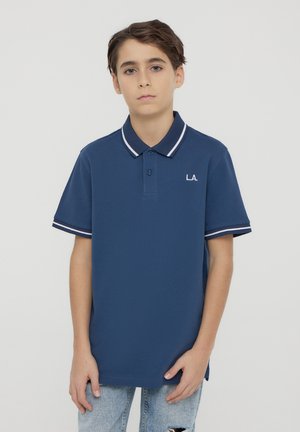Ragazzo giovane che indossa una polo blu navy con rifiniture bianche e "L.A." sul petto, abbinata a jeans azzurri strappati, in piedi davanti a uno sfondo semplice.
