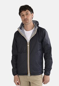 Homme aux cheveux courts et foncés portant une veste à capuche noire sur une chemise blanche et un pantalon beige, debout devant un fond uni.