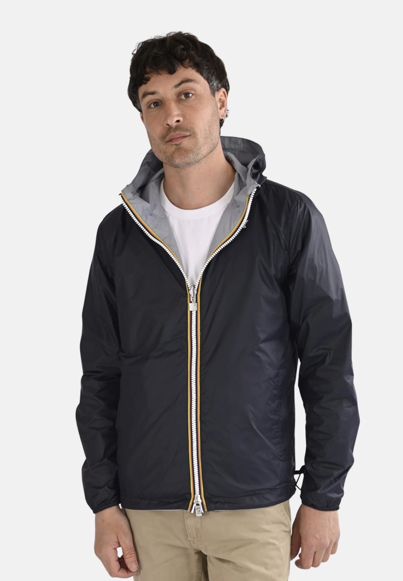 Homme aux cheveux courts et foncés portant une veste à capuche noire sur une chemise blanche et un pantalon beige, debout devant un fond uni.