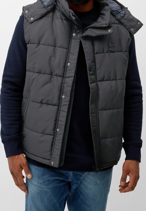 Homme portant une veste noire matelassée ouverte avec des boutons-pression sur un sweat-shirt bleu marine et un jean bleu, debout devant un fond uni.