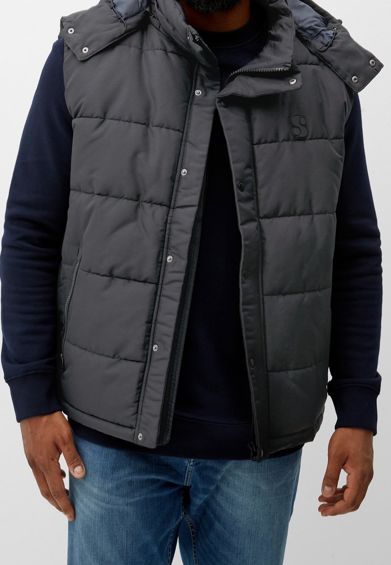Homme portant une veste noire matelassée ouverte avec des boutons-pression sur un sweat-shirt bleu marine et un jean bleu, debout devant un fond uni.