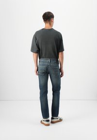 Camiseta gris oscuro de manga corta combinada con unos vaqueros azules que tienen bolsillos traseros. Zapatillas blancas con detalles en negro y suelas marrones.
