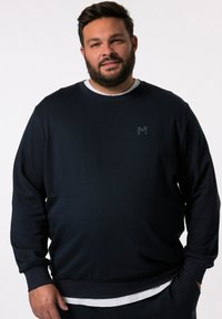 Felpa blu navy con maniche lunghe, texture a coste, collo a giro e un logo discreto sul petto. Presenta una camicia bianca sovrapposta sotto.
