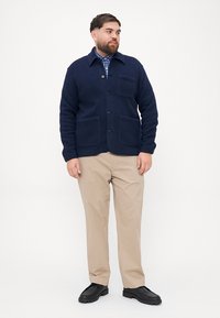 Ναυτικό πουλόβερ από μείγμα μάλλινου υφάσματος με γιακά, μπροστινά κουμπιά και δύο μεγάλα τσέπες. Φοριέται πάνω από πουκάμισο με καρό σχέδιο, με μπεζ παντελόνι και μαύρα παπούτσια.