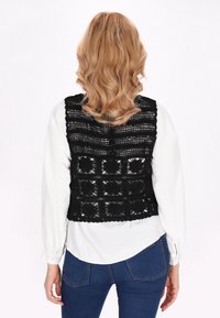 Črn kvačkan žaket z geometrijskim vzorcem, odprtimi hrbtom, nosi se čez belo srajco z dolgimi rokavi, v kombinaciji z modrimi ožjimi jeans hlačami.