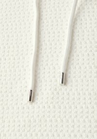 Tissu blanc texturé avec un design en maille, comportant des cordons blancs avec des embouts en argent et des finitions délicates sur le matériau.