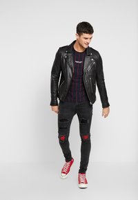 Μαύρο δερμάτινο μπουφάν μηχανής πάνω από ναυρικό μπλουζάκι με ρίγες, συνδυασμένο με φθαρμένα μαύρα τζιν παντελόνια με κόκκινα λουλουδάτα μοτίβα και κόκκινες αθλητικές επιγονατίδες.