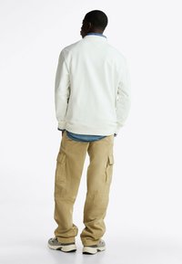 Weißer Sweatshirt mit strukturiertem Finish, über einem blauen Jeanshemd getragen. Kombiniert mit beigen Cargo-Hosen mit Seitentaschen und lockerem Schnitt.