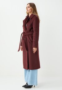 Cappotto in misto lana borgogna con cintura, design doppiopetto e grandi bottoni, abbinato a jeans in denim blu chiaro a gamba larga e tacchi neri.
