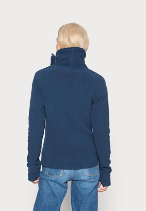 Personne blonde portant une veste polaire bleu marine et un jean bleu, vue de dos sur un fond clair uni.