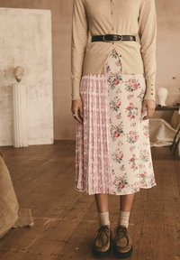 Blumenmusterrock mit plissierten Abschnitten in Rosa und Creme, kombiniert mit einer beigen Strickjacke und einem schwarzen Gürtel. Braune Schuhe runden das Outfit ab.