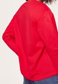 Punainen pitkähihainen paita rennossa mitoituksessa ja gathered-hihansuilla; pehmeää puuvillakangasta; pyöreä pääntie; yhdistettynä sinisiin farkkuihin, näkyy takanäkymä.