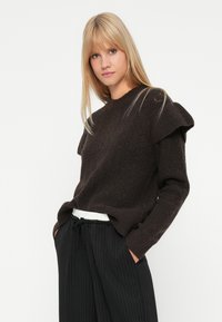 Kobieta z prostymi blond włosami, ubrana w strukturalny, ciemno-brązowy sweter z detalami na ramionach oraz w czarne spodnie w paski z kieszeniami. 