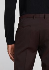 Pantaloni marroni su misura con una texture liscia, dotati di una tasca posteriore elegante e una vita aderente, abbinati a un maglione nero.