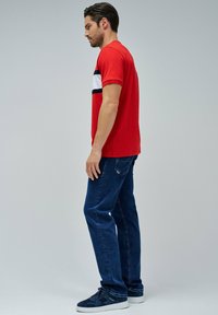 Camiseta roja de cuello redondo con rayas horizontales blancas y azul marino en el pecho, combinada con jeans de mezclilla azul oscuro y zapatillas azul marino.