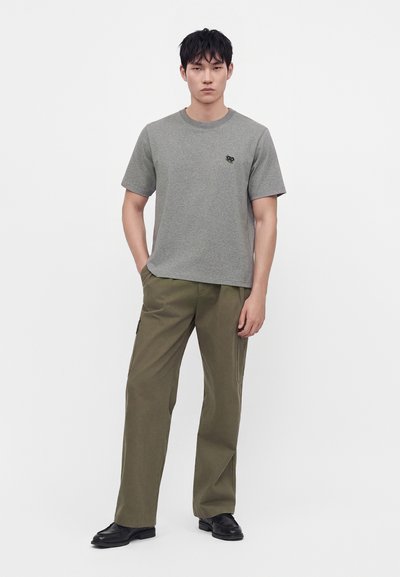Homme debout portant un t-shirt gris à manches courtes avec logo, pantalon large vert olive et chaussures noires sur un fond blanc uni.