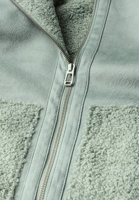 Giacca zip-up verde chiaro realizzata in materiale liscio con pannelli in texture pelosa, dotata di una cerniera argentata e di un morbido rivestimento interno.