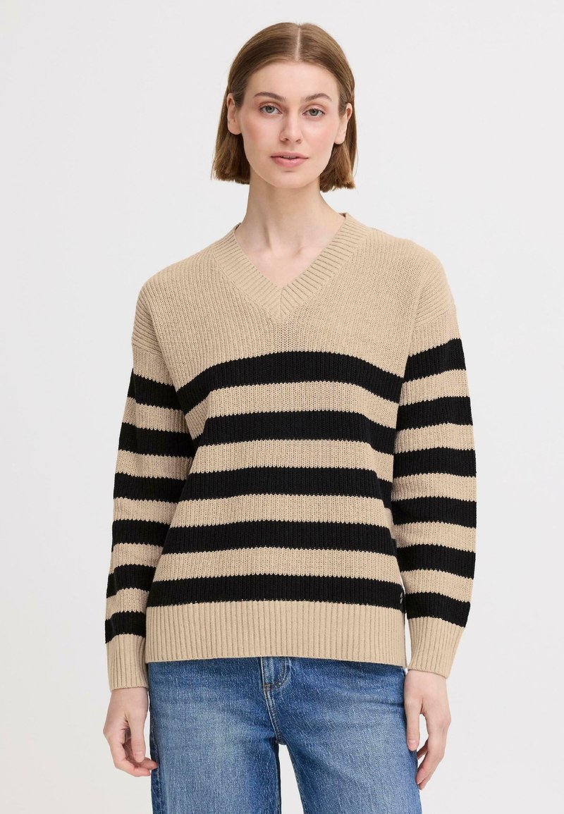 Beżowy sweter z czarnymi poziomymi paskami, dekolt w serek, ściągacze przy mankietach i dolnym brzegu, materiał dziany o fakturze, noszony z niebieskimi dżinsami.