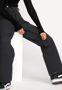 Pantalons de ski noirs avec une doublure intérieure texturée, des vents latéraux zippés et un design de genoux renforcé. Associés à des chaussures noires avec des accents blancs.