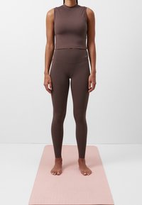 Brun crop top och högmidjade leggings i stretchigt material, placerade på en rosa yogamatta. Logotypen är tryckt i vitt.