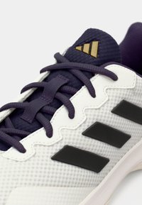 Weißer Sportschuh mit strukturierter Oberfläche, lila Akzenten, schwarzen Streifen und einem goldenen Logo auf der Zunge. Verfügt über ein Schnürdesign.