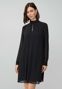 Vrouw gekleed in een zwart lange mouwen jurk met doorschijnende mouwen en een hoge halslijn met een kleine split aan de voorkant, staand tegen een neutrale lichte achtergrond.