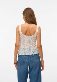 Top en dentelle blanche avec un motif floral transparent et des bords festonnés, associé à un jean en denim bleu taille haute, vu de dos.