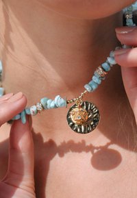 Collier en perles avec des pierres bleu clair, des accents dorés, et un pendentif avec un design noir texturé et un détail scintillant.