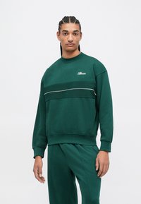 Jovem com cabelo trançado, vestindo um moletom escuro verde da Ellesse e calças combinando, de pé contra um fundo simples.