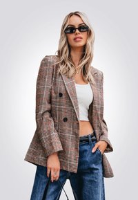 Blazer à carreaux brun et rouge à double boutonnage avec des revers à cran, porté sur un crop top blanc, associé à un jean bleu taille haute.